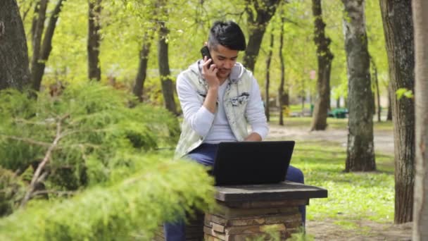 Asian man with laptop in park speaking on phone — Stock Video