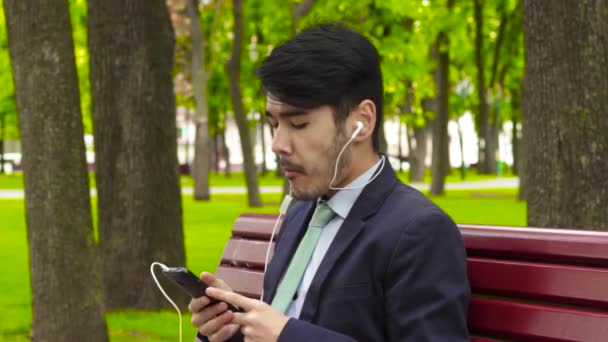 Азиатский бизнесмен сидит в парке и ест картошку фри в наушниках — стоковое видео