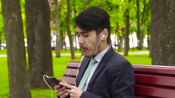 Asiático empresário sentado no parque e comer batata frita em fones de ouvido — Vídeo de Stock