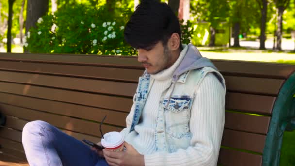 Мужчина сидит в парке и пьет кофе — стоковое видео