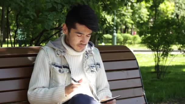 Man student zitten in het park en het schrijven van notitieblok — Stockvideo