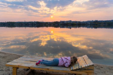 Küçük kız şezlonga uzanıp gün batımını seyrediyor.