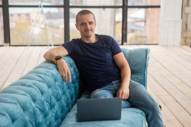 Dizüstü bilgisayarlı bir adam kanepede oturuyor, kameraya bakıyor ve gülümsüyor..