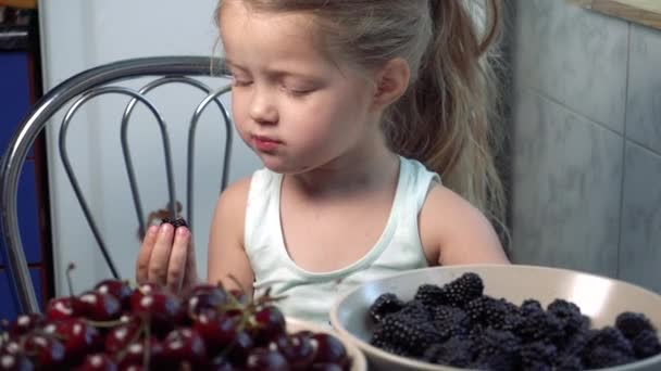 Küçük tatlı kız mutfakta oturmuş böğürtlen yiyor. — Stok video