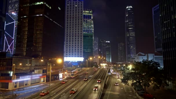 Гонконг, Китай. Вечернее движение машин с небоскребами на заднем плане. Автомобили и автобусы мчатся к месту назначения, проезжая мимо небоскребов делового района Гонконга. 4K — стоковое видео