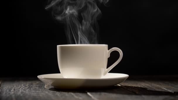 Cup of hot coffee. Steam coming out of a coffee cup standing on a white saucer on a wooden table. Close-up shot with a black background, UHD — Stock Video