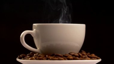 Sıcak içecek bir fincan tabağı üzerinde duran fincan kahverengi kavrulmuş kahve çekirdekleri ile dolu. Gri buhar bardaktan geliyor. Siyah arka plan, ağır çekim vurdu