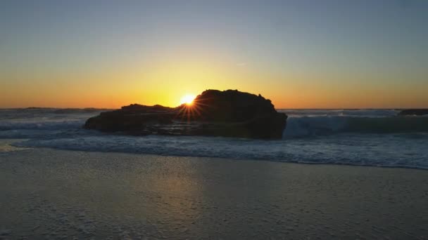 Nueve rayos de sol naranja por encima de la roca negra en la costa Antártica de Portugal. Las olas espumosas inundan la playa de arena mojada, la dejan cubierta de burbujas. UHD — Vídeos de Stock