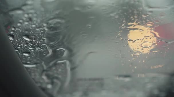 Essuie-glaces d'un pare-brise de voiture fonctionnant sous la pluie. Les feux brouillés de la circulation automobile de passage passent à travers le pare-brise. L'eau coule sur le pare-brise. UHD 