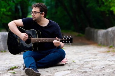 Gözlüklü genç adam eski Roma köprüsünde oturmuş gitar çalıyor.