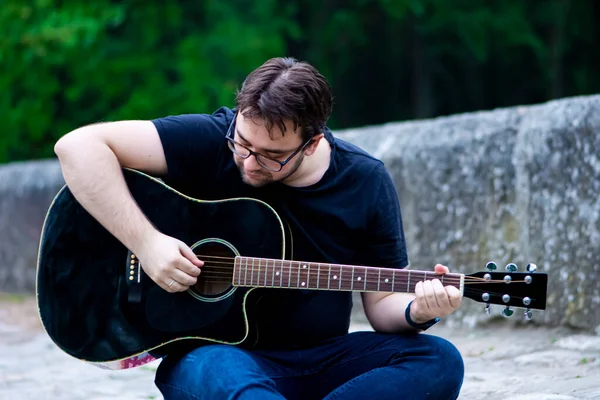 Young man wearing glasses playing guitar sitting on the old Roman bridge
