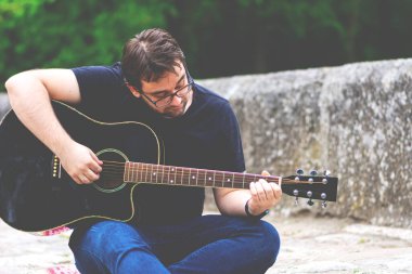 Gözlüklü genç adam eski Roma köprüsünde oturmuş gitar çalıyor.