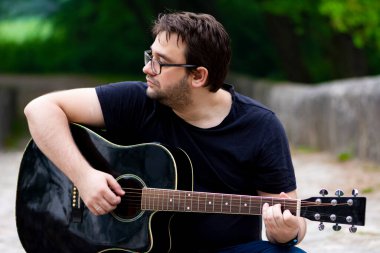 Gözlüklü genç adam eski Roma köprüsünde oturmuş gitar çalıyor.