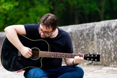Gözlüklü genç adam eski Roma köprüsünde oturmuş gitar çalıyor.