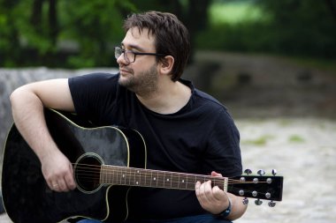 Gözlüklü genç adam eski Roma köprüsünde oturmuş gitar çalıyor.