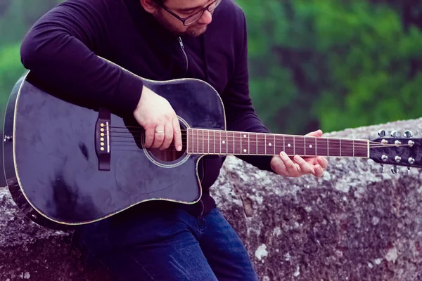 Young man wearing glasses playing guitar leaning against the old Roman bridge