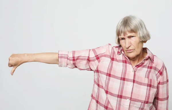 Serious leeftijd vrouw wijzende vinger naar beneden. Portret van expressieve grootmoeder met roze shirt. Studio opname geïsoleerd op lichtgrijze achtergrond. — Stockfoto