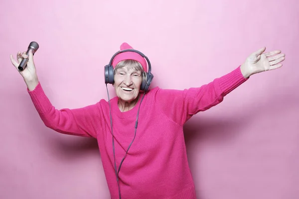 Levensstijl en mensen concept: Funny oude dame luisteren muziek met koptelefoon en zingen met mic op roze achtergrond — Stockfoto