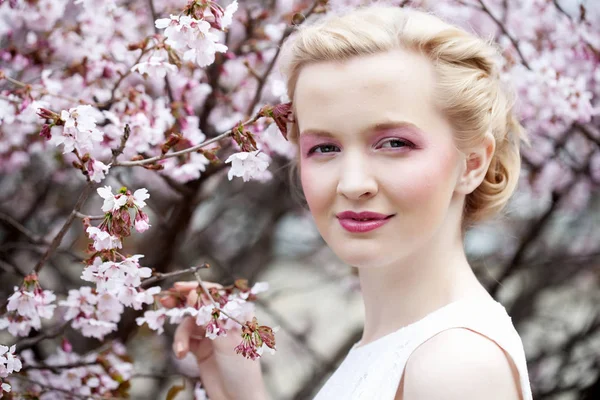 Retrato de uma bela jovem loira em um fundo de flores de cereja rosa na primavera — Fotografia de Stock