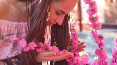 Genç casual giyinen kız bardaklarda sakura çiçekler ve gülümseyerek kameraya kokuyor. Bahar kavramı yüzüne odaklan. Ağır çekim.