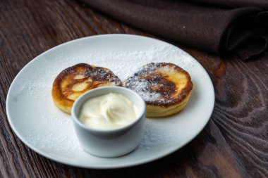 Peynirli kekler beyaz tabakta. Peynirli keklerin yanında bir fincan ekşi krema ve bir kaşık var. Peynirli kekler taze nane yaprağıyla süslenir. Yukarıdan bak. Yakın plan. Makro fotoğrafçılık.