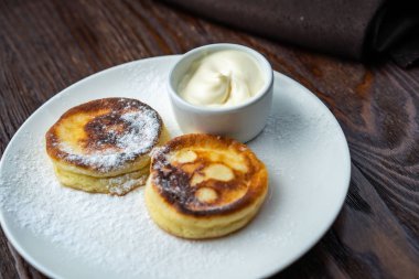 Peynirli kekler beyaz tabakta. Peynirli keklerin yanında bir fincan ekşi krema ve bir kaşık var. Peynirli kekler taze nane yaprağıyla süslenir. Yukarıdan bak. Yakın plan. Makro fotoğrafçılık.