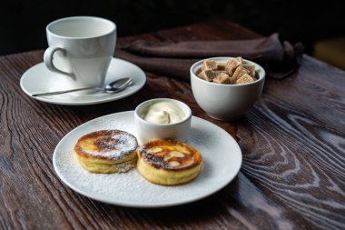 Peynirli kekler beyaz tabakta. Peynirli keklerin yanında bir fincan ekşi krema ve bir kaşık var. Peynirli kekler taze nane yaprağıyla süslenir. Yukarıdan bak. Yakın plan. Makro fotoğrafçılık.