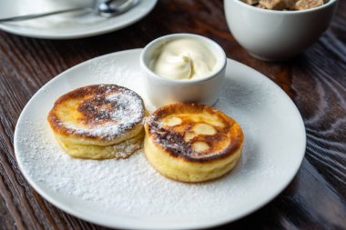 Peynirli kekler beyaz tabakta. Peynirli keklerin yanında bir fincan ekşi krema ve bir kaşık var. Peynirli kekler taze nane yaprağıyla süslenir. Yukarıdan bak. Yakın plan. Makro fotoğrafçılık.