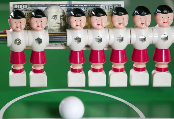 Sports and money concept -toy football players stand in the field and the banknote in hundred dollars on a backgroun