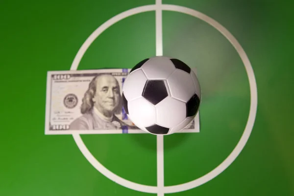 Sports and money concept - Toy soccer ball in a midfield, in the center of the green field and the banknote in hundred dollars on a backgroun