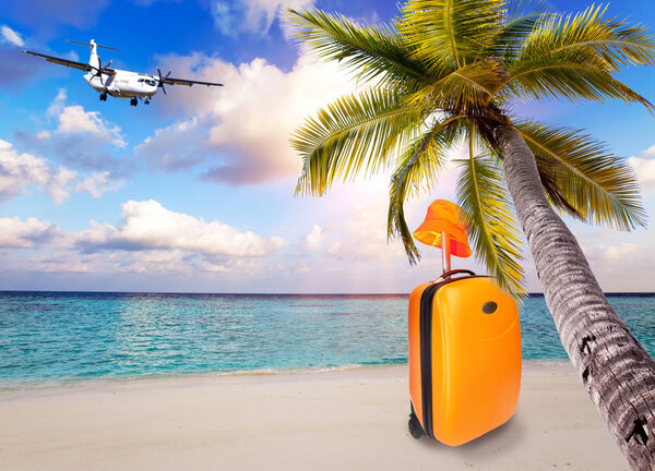 Bright orange suitcase and hat on the sandy beach by the sea under an inclined palm tree and the plane in the sky with clouds