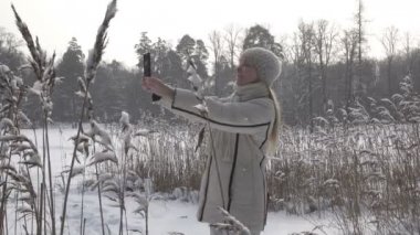 Genç kadın bir beyaz ceket fotoğraflarda bir kış orman Gölü kıyısında smartphone Panoraması, kar yağıyor