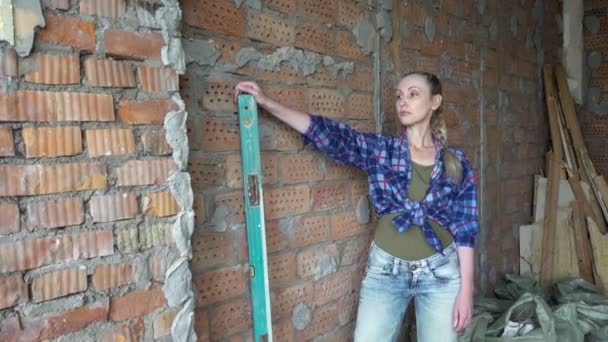 Η νεαρή λεπτή γυναίκα με καρό πουκάμισο και μπλε τζιν στο φόντο του τοίχου να πρόκειται να κάνει μετρήσεις ενός κόκκινου τούβλου από το επίπεδο κατασκευής. Κατασκευαστικές εργασίες. Ανακαίνιση διαμερίσματος — Αρχείο Βίντεο