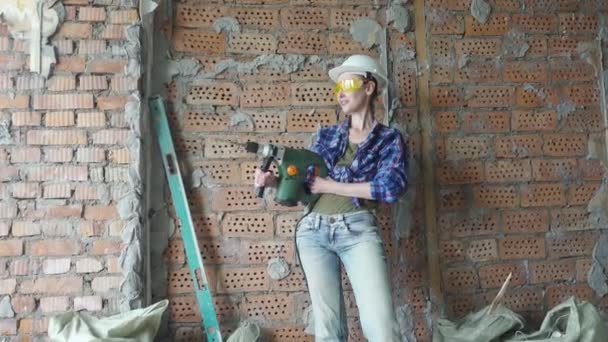 A jovem mulher em roupas de trabalho e um capacete de construção com o puncher contra o fundo de campos de tijolos vermelhos, obras de construção. Renovação do apartamento — Vídeo de Stock