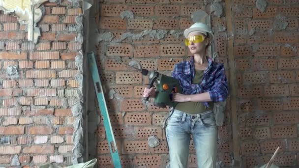 Молодая женщина в рабочей одежде и строительном шлеме с перфоратором на фоне красных кирпичных лагерей, строительно-монтажные работы. Ремонт квартир — стоковое видео