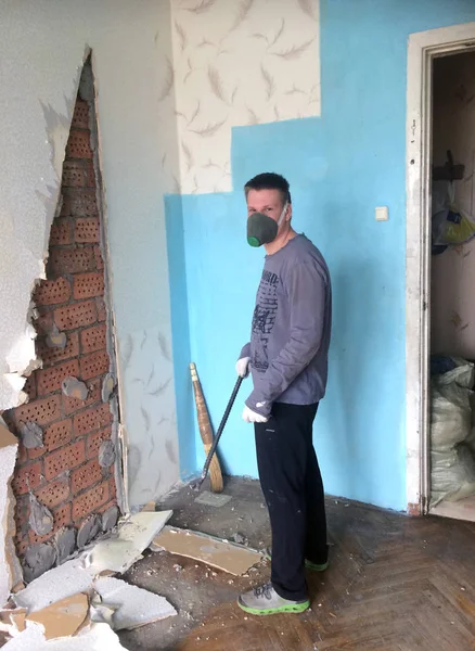 repair in the home. A young man knocks down old Soviet-era plasterboard slabs from the wall with iron scrap. Visible brick wall base