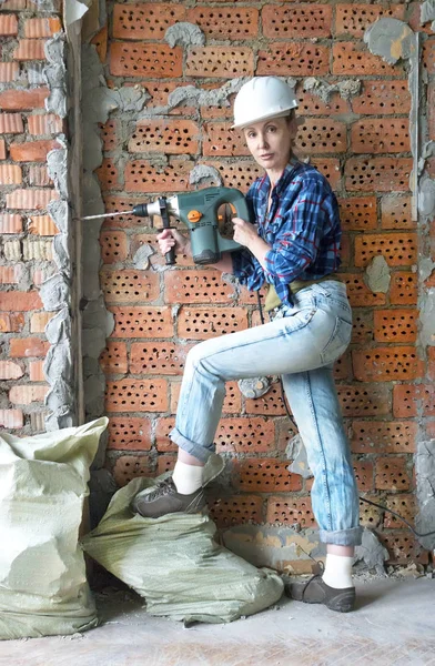 young woman in work clothes and a protective helmet stands next to a brick wall and makes a hole with a heavy hammer drill