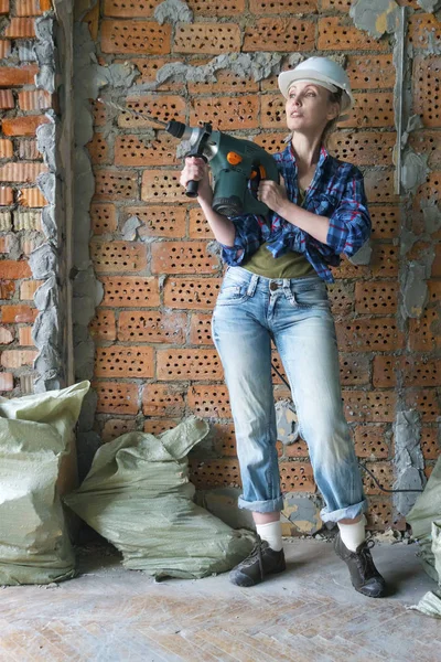 作業服と保護ヘルメットを着た若い女性はレンガの壁の隣に立ち 重いハンマードリルを保持しています — ストック写真
