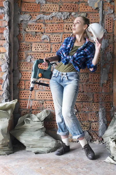 疲惫的年轻女子在工作服与保护头盔和沉重的锤钻站在砖墙附近 — 图库照片