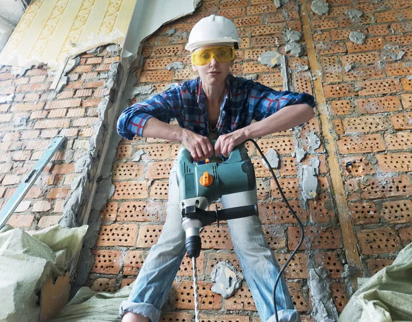 young woman in work clothes with a protective helmet and a heavy hammer drill stands near a brick