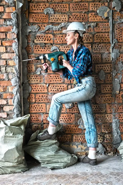 young woman in work clothes and a protective helmet stands next to a brick wall and makes a hole with a heavy hammer drill