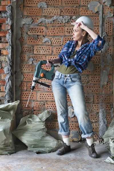 疲惫的年轻女子穿着工作服 戴着防护头盔 站在砖墙前 用沉重的锤子钻 擦去额头上的汗水 — 图库照片