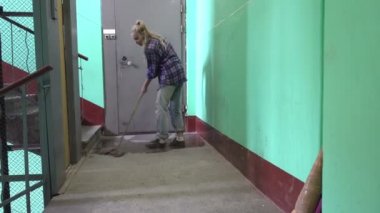 Kot pantolonlu ve kafes gömlekli genç bir kadın Stalinist bir apartmanın asansörünün yanındaki merdivenlerde sallanıyor ve yerleri yıkıyor işçi göçmeni bir temizlikçinin yanında kamu alanlarını temizliyor.