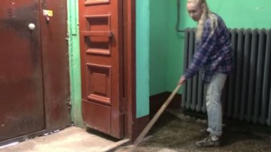 Kot pantolonlu ve kafes gömlekli genç bir kadın Stalinist bir apartmanın asansörünün yanındaki merdivenlerde sallanıyor ve yerleri yıkıyor işçi göçmeni bir temizlikçinin yanında kamu alanlarını temizliyor.