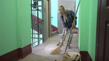 Kot pantolonlu ve kafes gömlekli genç bir kadın Stalinist bir apartmanın asansörünün yanındaki merdivenlerde sallanıyor ve yerleri yıkıyor işçi göçmeni bir temizlikçinin yanında kamu alanlarını temizliyor.