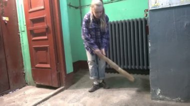 Kot pantolonlu ve kafes gömlekli genç bir kadın Stalinist bir apartmanın asansörünün yanındaki merdivenlerde sallanıyor ve yerleri yıkıyor işçi göçmeni bir temizlikçinin yanında kamu alanlarını temizliyor.