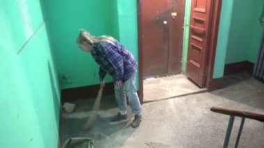 Kot pantolonlu ve kafes gömlekli genç bir kadın Stalinist bir apartmanın asansörünün yanındaki merdivenlerde sallanıyor ve yerleri yıkıyor işçi göçmeni bir temizlikçinin yanında kamu alanlarını temizliyor.