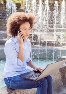Güzel genç Afro Amerikan öğrenci kadın Laptop ile çalışma gözlüklü ve telefonla konuşma. Açık portre, rahat kıyafet 