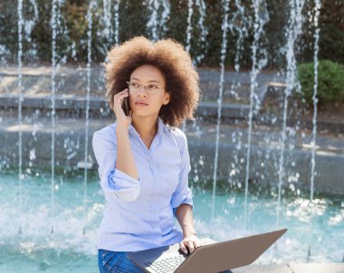 Güzel genç Afro Amerikan işkadını Laptop ile çalışma gözlüklü ve telefonla konuşma. Açık portre, rahat kıyafet 