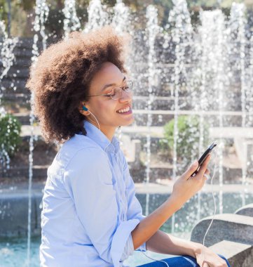 Sakin ve yaz günü, Çeşme, kulaklık ile müzik dinleme yakın oturan rahat kıyafet güzel genç Afro-Amerikan kadın zevk